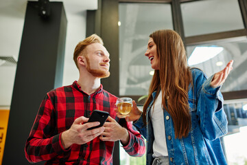 Cheerful male and female friends 20 years old smiling while discussing online blogging via cellular gadget, Caucasian hipster guys with smartphone gadget refreshing and talking in cafe interior