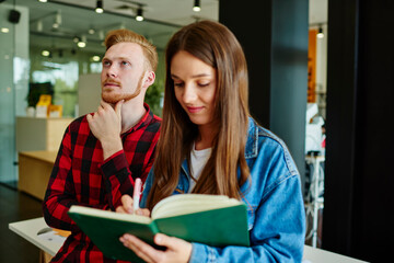 Caucasian male and female students 20s cooperating on organisation plan during together education, youthful hipster guys with knowledge notepad making information notes during indoors studying