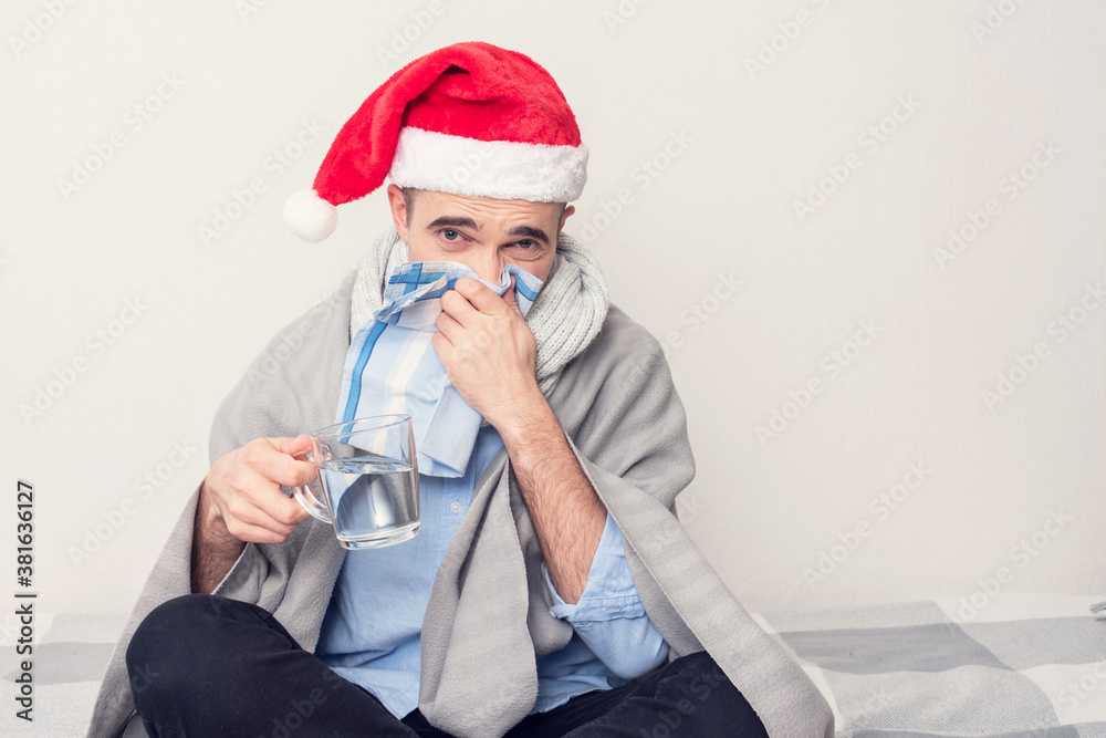 Wall mural Santa with a handkerchief and a glass of water is ill with the flu, a man is sitting at home on self-isolation, portrait, white background