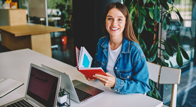 Portrait of cheerful Caucasian freelancer with education textbook sitting at table desktop with laptop computer, successful female student smiling at camera while e learning in coworking space