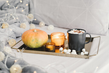 Fall season breakfast: hot chocolate with marshmallow in a black mug, burning candles and pumpkin on a metal tray served in bed with decorative lights. Autumn inspiration