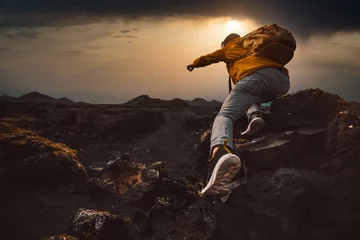 Rolgordijnen Successful hiker hiking a mountain pointing to the sunset. Wild man with backpack climbing a rock over the storm. Success, wanderlust and sport concept. © Davide Angelini
