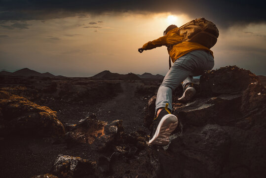 Successful hiker hiking a mountain pointing to the sunset. Wild man with backpack climbing a rock over the storm. Success, wanderlust and sport concept.