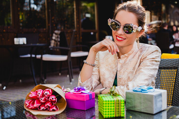 Obraz premium young stylish beautiful woman in fashion sunglasses sitting at cafe, elegant, smiling and happy, enjoying warm day, presents, tulips, happy birthday party, city street, europe vacation