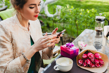 young stylish woman sitting in cafe, holding smart phone, drinking cup cappuccino, enjoying, tulips, celebrating party, city street, boho outfit, europe vacation, romantic dinner, smiling, presents
