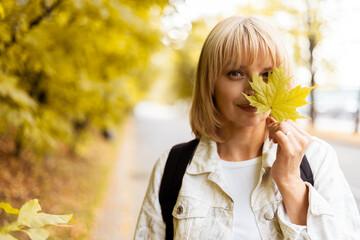 Portrait of happy blonde adult woman with a smile holds an autumn yellow maple leaf near the face in the park on the nature walk outdoors