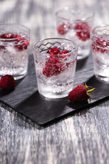 Alcohol shots of berries cocktail with a raspberry on black stone tray. Glass of sparkling water on wooden table