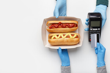 Courier in rubber gloves gives customer cardboard box with hot dogs with grilled sausages, with...