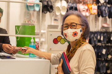 Happy senior customer wearing protective mask paying money at cashier counter in shoes shop.