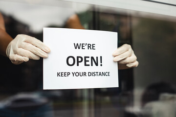We are open keep your distance. Hands of master in rubber gloves hang notice on glass door of manicure salon after coronavirus quarantine