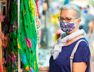 Obraz premium Elderly woman wearing a medical mask due to coronavirus while shopping at the flea market. Concept of buying cheap used clothes