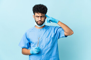 Moroccan dentist man holding tools isolated on blue background having doubts and thinking