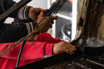 Expert senior mechanic teaching a young prospect how to repair a car after a street crash. Details and focus on hands while working on metal parts and electric wires. Blurred background, natural light