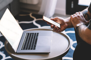 Crop man reading information from smartphone while sitting at table with laptop