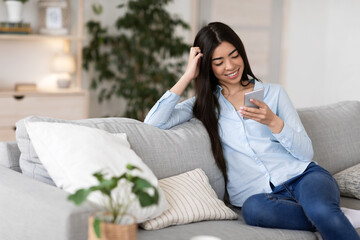 Joyful korean woman resting on couch in living room with smartphone