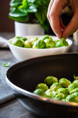 A hand picking up fresh brussels sprouts from a pan with a white bowl and plant decor in the background