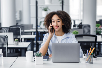 Obraz premium Return to workplace after quarantine. Friendly african american woman talking on phone, sitting at table with laptop in modern office interior with antiseptic