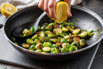 A hand squeezes lemon juice over roasted / fried brussels sprouts in a black frying pan that sits on a cutting board with a solid spoon for serving. Half a lemon and towel in the background