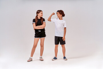 Two teenagers exercising together. Boy showing his biceps to a cute girl, they standing isolated over grey background in studio