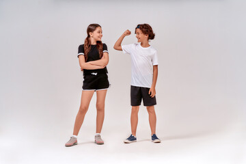 Obraz premium Two teenagers exercising together. Boy showing his biceps to a cute girl, they standing isolated over grey background in studio