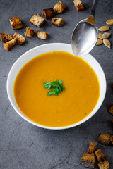 Pumpkin soup with coriander in a white bowl with a spoon coming towards. Croutons and pumpkin seeds in the background. Seasonal autumn / winter food, healthy and fresh
