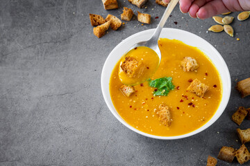 Woman hand taking a spoon of pumpkin soup while the bowl is behind with coriander, gluten free...