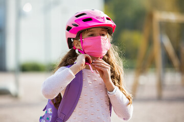 Fototapeta na wymiar Schoolgirl wearing protective fabric reusable face mask riding a bike to school. School education during the pandemic. Security measures and social distancing