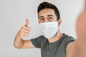 Man wearing face mask taking a selfie while doing ok sign with his free hand. White background.