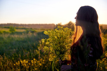 Fototapeta premium Beautiful woman in red dress holding bouquet of yellow flowers and enjoying summer sunny day in green field with green grass in sunset with nice orange sun