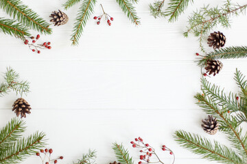 Fototapeta na wymiar Christmas frame of fir branches, red berries and pine cones on white wood, minimalist flat lay