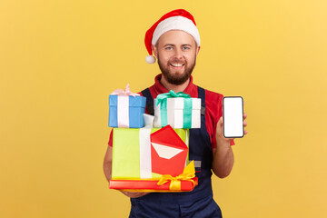 Optimistic deliveryman in uniform and red santa claus hat holding holidays presents and showing smartphone with blank display, order and delivery app. Indoor studio shot isolated on yellow background