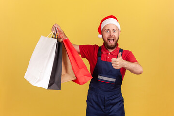 Happy excited male courier in uniform and santa claus hat showing thumbs up holding paper shopping bags, likes express delivery. Indoor studio shot isolated on yellow background
