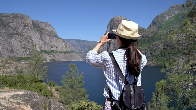 back view of asian young girl backpacker traveler using smart phone taking picture camera app of hetch hetchy valley in yosemite national park. woman hiker with bag and straw hat standing by lake.