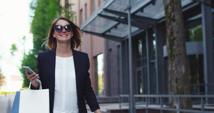 Portrait of happy Caucasian businesswoman in sunglasses walking on street with purchase after shopping. Stylish adult female worker in formal suit using smartphone, holding packages, paper bags.