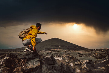 Successful hiker hiking a mountain pointing to the sunset. Wild man with backpack climbing a rock...