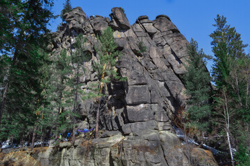Large high rough climbing rock Vityaz in Siberian forest with conifers in winter in the light of sun. People climbers on big gray cliff. Blue sky. Russia
