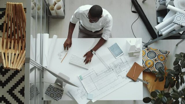 Top down shot of African American architect in formal outfit typing on computer while working at office desk with paper floor plan on it