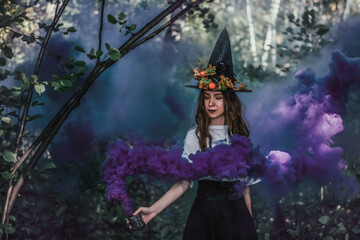 Young pretty witch in black hat and with purple fog in her hand close up in the forest.