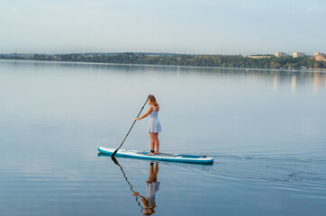 Woman in a dress on a sap board floats on the blue water of the lake in the evening