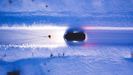 Aerial top view of car on rural area road while headlights are on in winter darkness, bird's eye view of suv vehicle in snowy north lands. Person standing front automobile which lighting the way