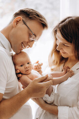 Happy young parents with newborn baby girl, loving mom hold little daughter in arms, caring dad gently hold a hand of adorable baby girl, tender family moments, parenting concept