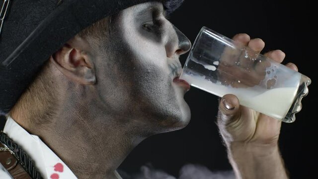 Creepy man with skeleton makeup in white shirt. Guy looking at camera, drinks milk from a glass