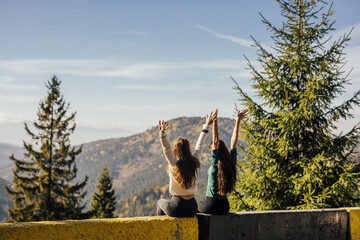 Two girlfriends with hands up admiring the view of the mountains and pines forest. They on top of the mountain watching at landscape. Back view. Copy space.