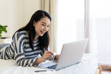 Happy young Asian woman using laptop computer on bed at home. Shopping online, Working from home, quarantined concept