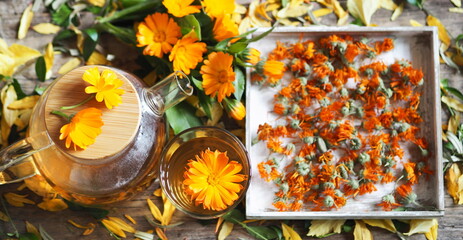 Medicinal autumn background. Herbal healthy marigold tea with a teapot, dried and fresh flowers on a wooden background with autumn leaves.