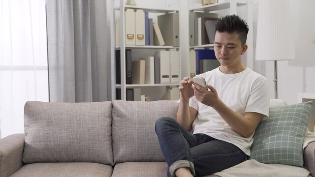 handsome asian male sitting comfortably in living room looking at social media feeds on his smartphone.