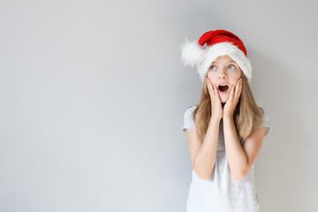 Portrait of a blonde caucasian surprised girl in a Santa hat on a grey background. Christmas and happines concept. Close up, copy space