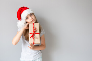 Portrait of a blonde caucasian  girl in a Santa hat holding the gift in her hands on a grey background. Christmas and happines concept. Close up, copy space