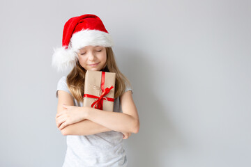 Portrait of a blonde caucasian smiling girl with closed eyes in a Santa hat holding the gift in her hands on a grey background. Christmas and happines concept. Close up, copy space