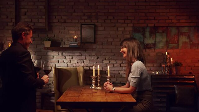 Elegant couple having romantic dinner at home, sitting at table in living room drinking red wine, clinking glasses, talking. Stay at home concept.